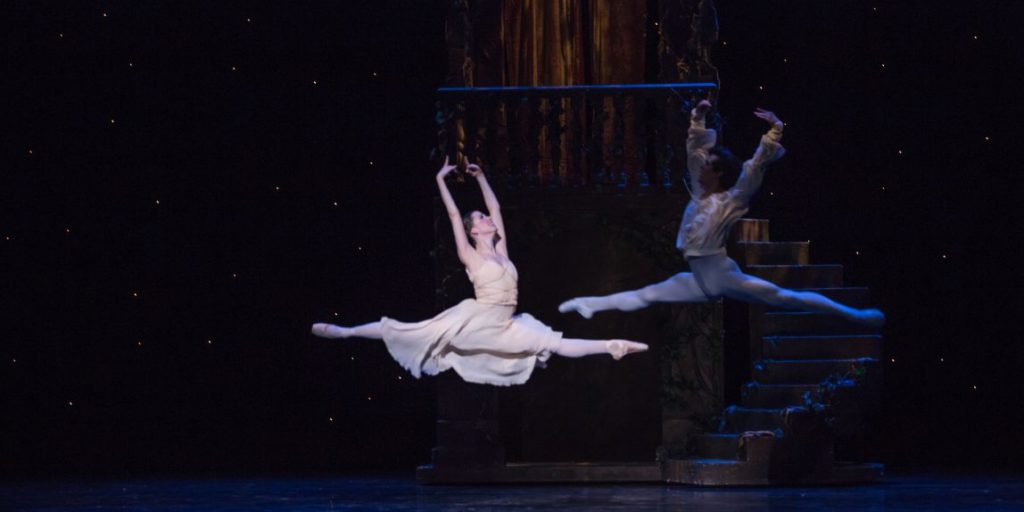 One male and one female ballet dancer in midair against a dark, starry stage set.