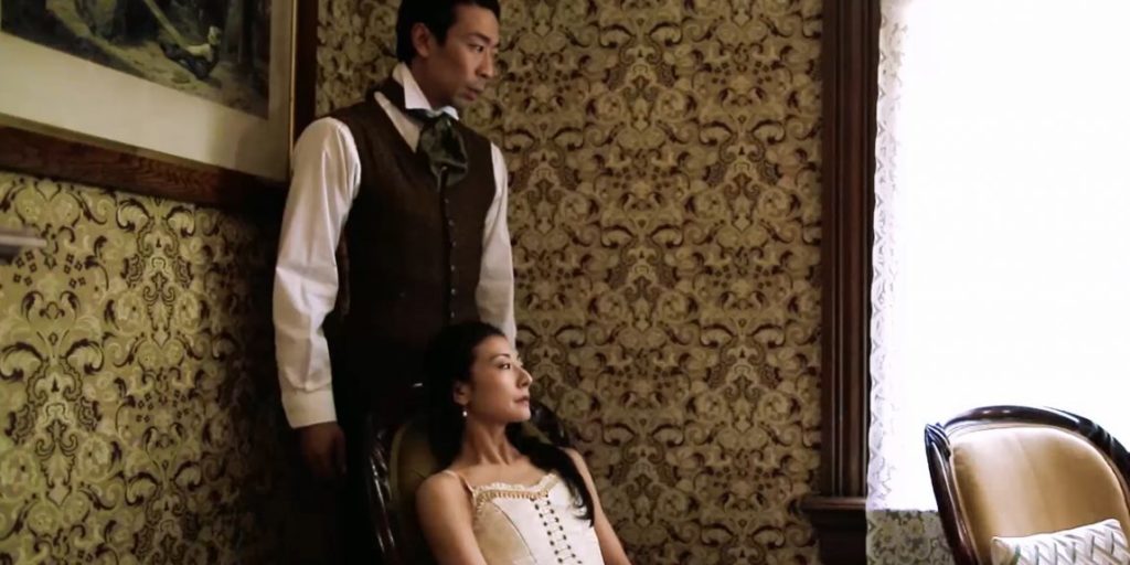 The cast of Vaulted: A man in a brown vest with his hand on the shoulder of a beautiful woman in a room with fancy wallpaper.