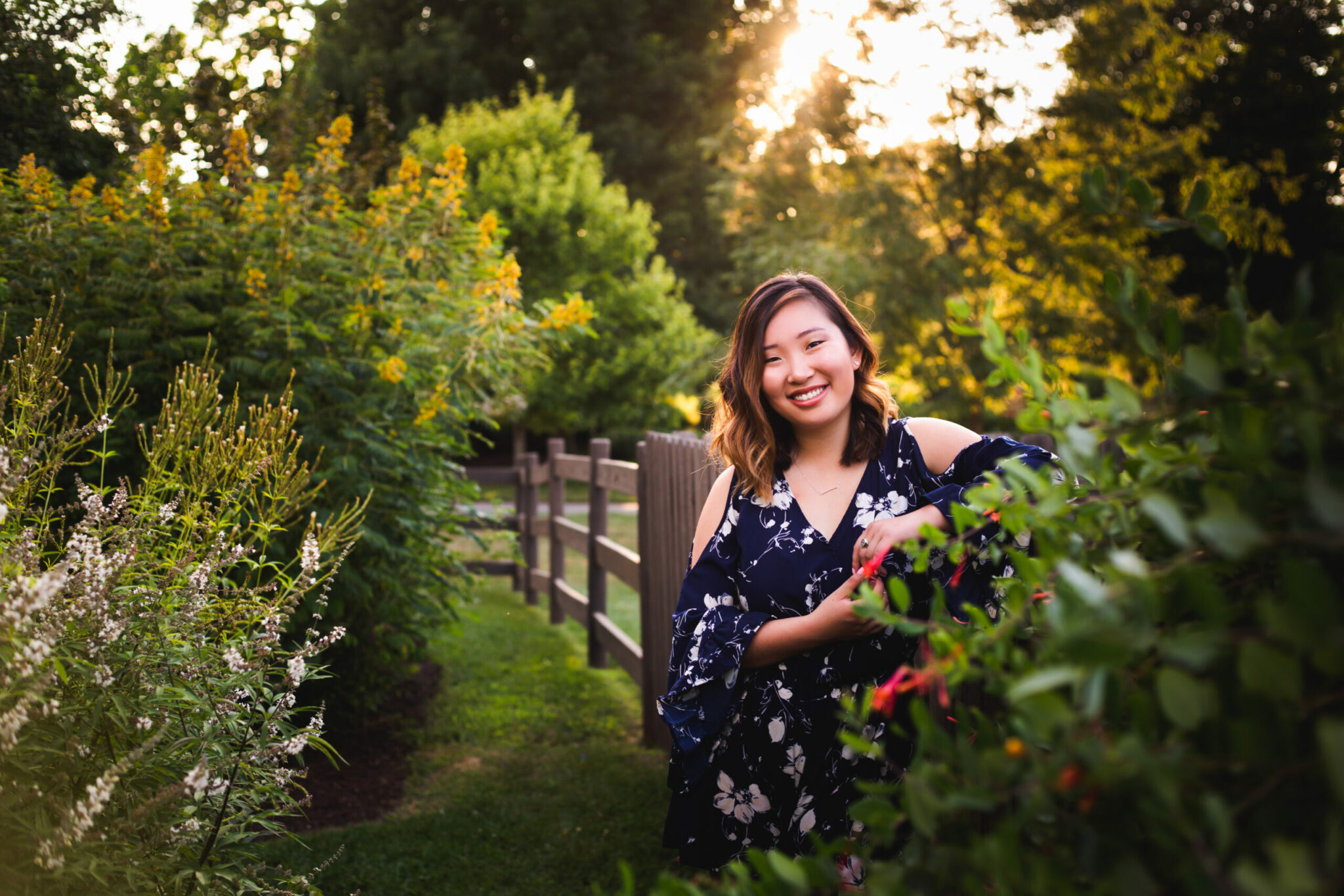 A smiling woman in a garden with pretty bushes and a fence.